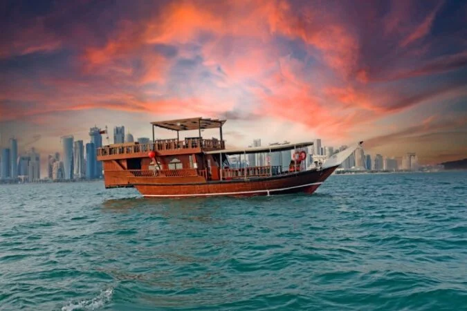 A traditional wooden boat sails on blue water with a modern city skyline in the background, under a dramatic, colorful sunset sky—perfect for a サンセットクルーズ experience.