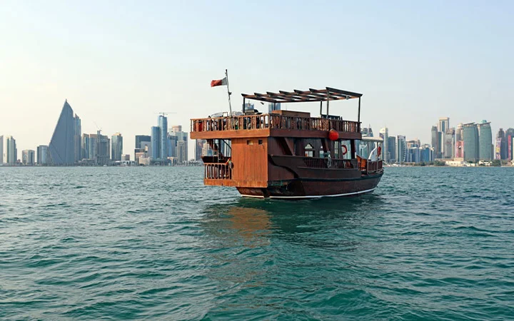A traditional wooden dhow boat sails on calm water with a modern city skyline, including the distinctive triangular WFCC building, visible in the background under a clear sky.