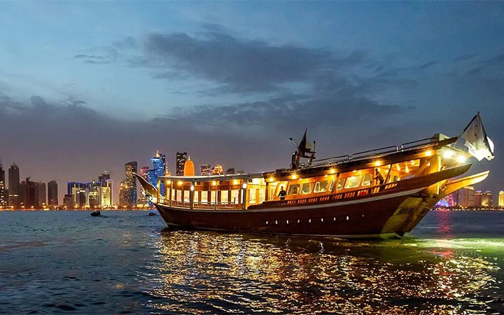 A traditional wooden dhow boat adorned with glowing lights sails on water at dusk, offering a memorable dinner cruise with the WFCC city skyline illuminated in the background under a partly cloudy sky.