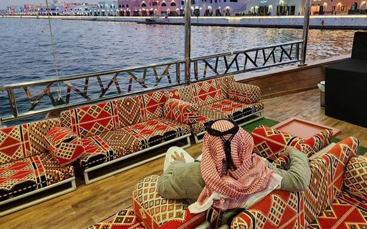 A person in traditional Middle Eastern attire relaxes on patterned cushions by the waterfront, gazing at a dhow boat gliding across calm waters with city lights shimmering in the distance.