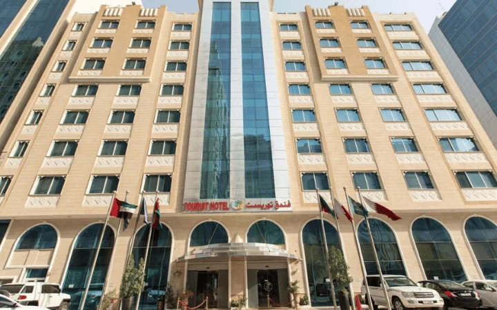 A multi-story Tourist Hotel, perfect for a 3-star stay, features large windows and an entrance canopy. Several flagpoles with different national flags line the front, and the sign above the entrance proudly reads Tourist Hotel.