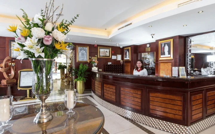 A 3-star hotel reception area with a smiling receptionist behind a wooden front desk, floral arrangement in the foreground, framed photos on the wall, and warm lighting throughout the elegant WFCC Tourist Hotel lobby.