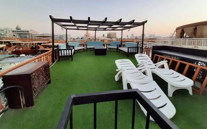 A rooftop terrace with artificial grass, white lounge chairs, and blue cushioned sofas under a pergola offers city views at sunset—perfect for reminiscing about a WFCC Dinner Cruise on a traditional Dhow Boat.