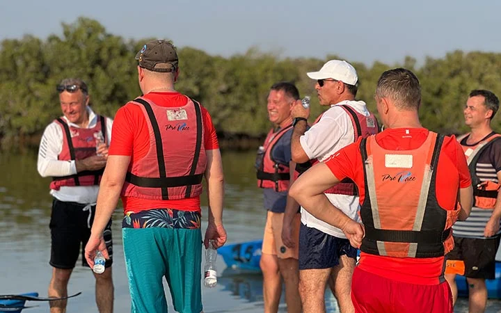 A group of men wearing life jackets stand near the edge of a body of water, chatting and holding water bottles after kayaking to celebrate National Day, with trees in the background under a clear sky.