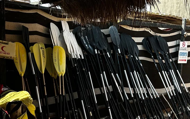 Several rows of black, gray, white, and yellow kayaking paddles are neatly stacked and leaning against a striped wall under a thatched roof. A first aid kit and safety signs are visible to the side—perfect for a National Day adventure.