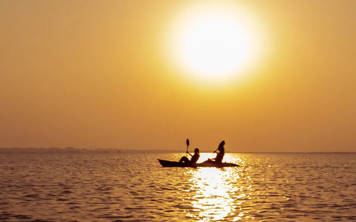 Two people kayaking on calm water at sunset, the sun casting a golden glow and reflections—an idyllic way to celebrate National Day surrounded by nature’s beauty.