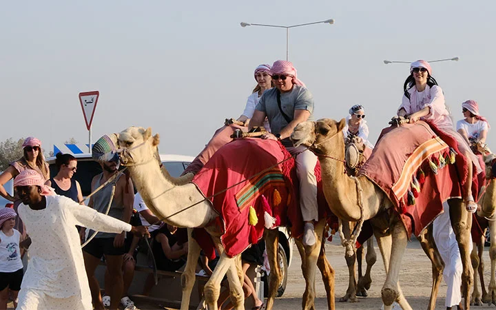 A group of tourists wearing headscarves ride camels led by a guide in traditional clothing on a sunny day, experiencing an exciting Desert Safari, with people and cars visible in the background.