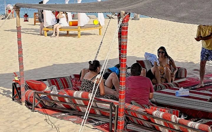People relax on patterned cushions under a canopy on a sandy beach, reminiscent of an overnight camping vibe. Two women and a man sit and chat, while another person stands nearby. In the background, sunbeds and white curtains overlook the water.