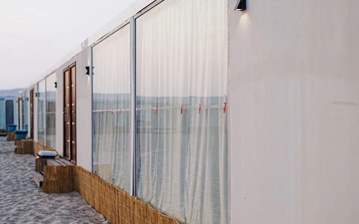 A row of white beach cabins with large windows covered by sheer curtains stands on sandy ground, perfect for overnight camping, with wooden benches and wicker panels along the base.