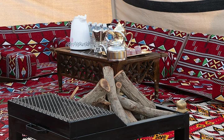 A traditional Middle Eastern seating area with patterned red cushions, a low wooden table holding a teapot, cups, water bottles, and a tray—perfect for relaxing after enjoying your Private Beach Setup in Qatar. Firewood and a metal grill complete the scene.