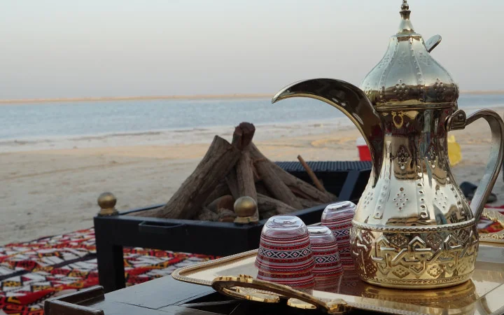 A traditional Arabic coffee pot with patterned cups sits on a tray near a firewood setup on a sandy Private Beach Setup in Qatar, with the sea and sky in the background.