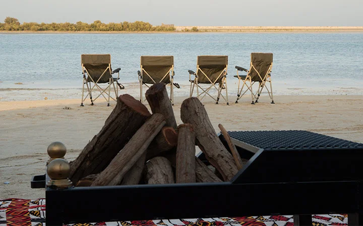 Four folding chairs face a calm body of water on a sandy beach, showcasing a cozy private beach setup in Qatar, with firewood and a grill in the foreground and trees lining the opposite shore.