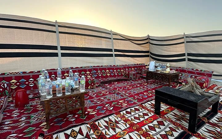 A traditional outdoor seating area with patterned red carpets and low tables offers a unique Private Beach Setup in Qatar, featuring bottled water, snacks, and fabric walls with black stripes, all set under a clear sky at sunset.