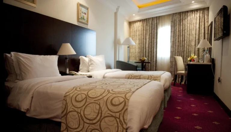 A cozy Musherib Hotel Qatar room with two neatly made single beds, white linens, beige patterned throws, bedside lamps, a window with curtains, a desk and chair, plus a wall-mounted television.