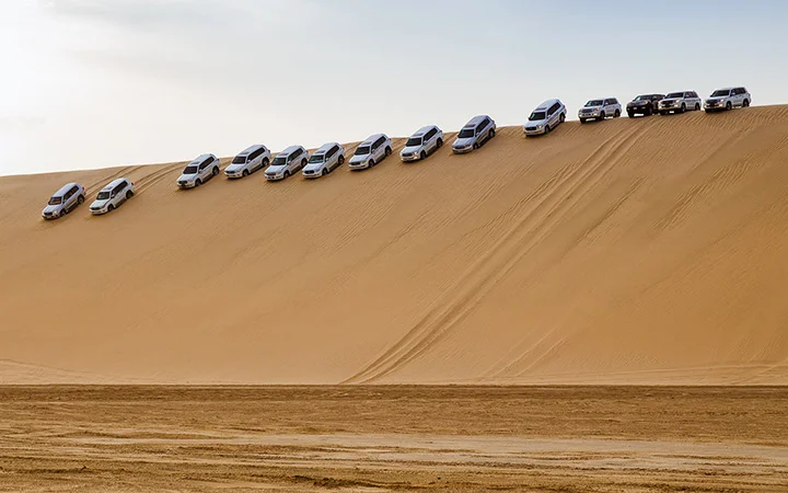 A row of SUVs is parked diagonally along the steep side of a sand dune in a desert, capturing the thrill of a Half Day Desert Safari, with tracks visible in the sand and a flat, open area in the foreground.