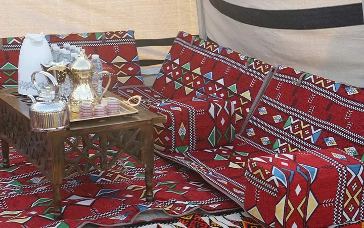 A traditional Arabic seating area with red patterned cushions and a wooden table set for Group Suhoor With Private Set-up, featuring Arabic coffee pots, teacups, and a tray for serving drinks inside a tent with woven fabric walls.