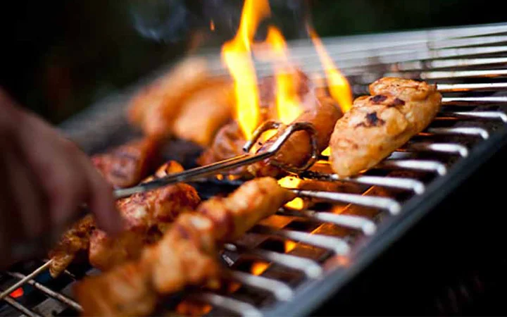 Pieces of chicken being grilled over an open flame on a barbecue, perfect for a group suhoor with private set-up, as metal tongs turn each juicy piece to perfection.