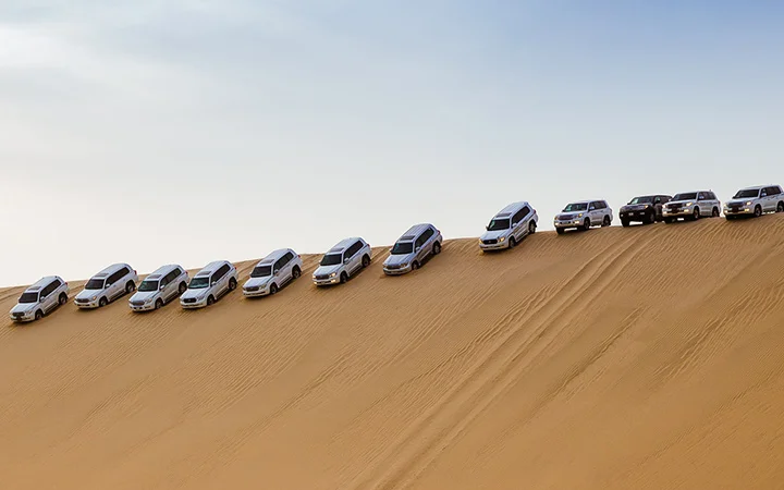 A row of SUVs and cars are parked diagonally on the steep slope of a sandy desert dune under a clear sky, ready for Iftar on the Dunes (per car) experience.