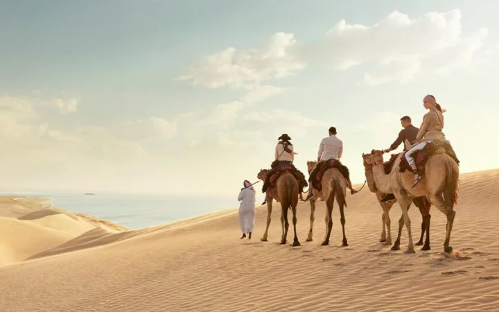 Four people ride camels led by a guide across golden sand dunes with the ocean in view, soaking in stunning scenery before enjoying Iftar on the Dunes (per car) under a partly cloudy sky.