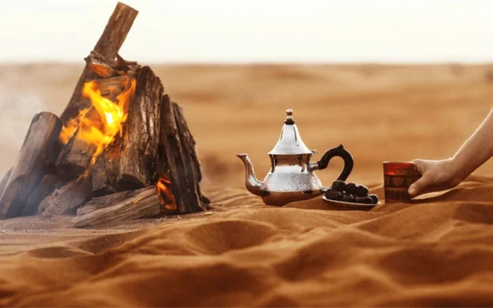 A metal teapot, a plate of dates, and a cup sit on sand near a campfire in a desert—capturing the essence of Iftar on the Dunes (per car), as a hand reaches for the cup against rolling dunes and a clear sky.