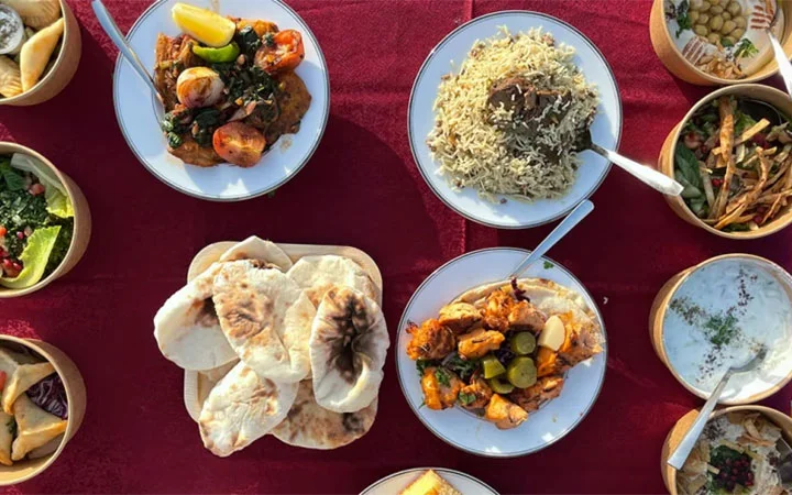 A table set for Iftar on the Dunes (per car), featuring plates of rice, grilled vegetables, curry dishes, naan bread, and assorted side salads, all beautifully arranged on a red tablecloth.