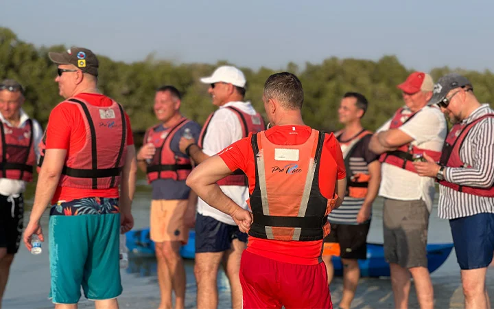 A group of men wearing life jackets gather outdoors near water, preparing for a kayaking session before Iftar. Some are talking while others check their phones, surrounded by trees and a clear blue sky.