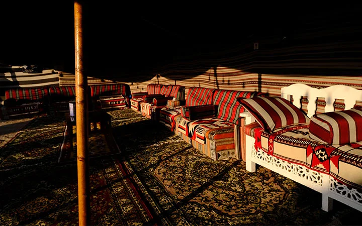 A traditional Bedouin tent interior with patterned rugs and colorful cushions on wooden benches, bathed in warm sunlight and shadow, offers a peaceful retreat after kayaking before gathering for Iftar.