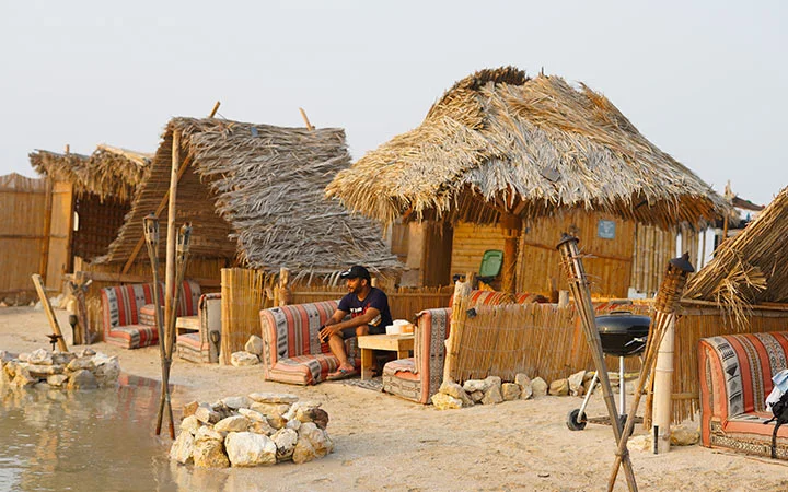 A person enjoys Iftar on a traditional cushioned seat under a palm-thatched hut by the water, with kayaks resting nearby, bamboo fences, and outdoor seating adding to the rustic beachside setting.