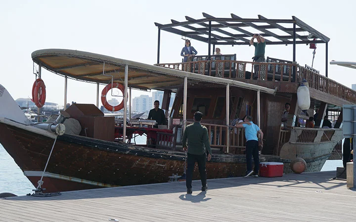 A group of people stand and work on a large wooden boat docked at a pier, enjoying a Private Dhow Boat Rental. Some are on the deck while others prepare near the dockside, with buildings visible in the distant background.
