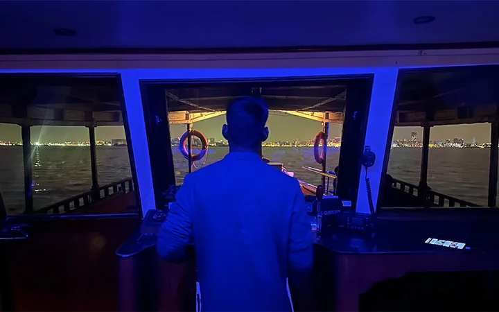 A person stands at the helm of a private dhow boat at night, gazing over dark water toward a brightly lit city skyline in the distance, illuminated by blue interior lights—a serene scene perfect for a Ramadan offer.