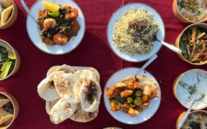 A top-down view of a table on a Private Yacht with Iftar Cruise, featuring Middle Eastern dishes like rice, grilled meats, naan bread, vegetables, salad, and yogurt—all beautifully arranged on a red tablecloth.