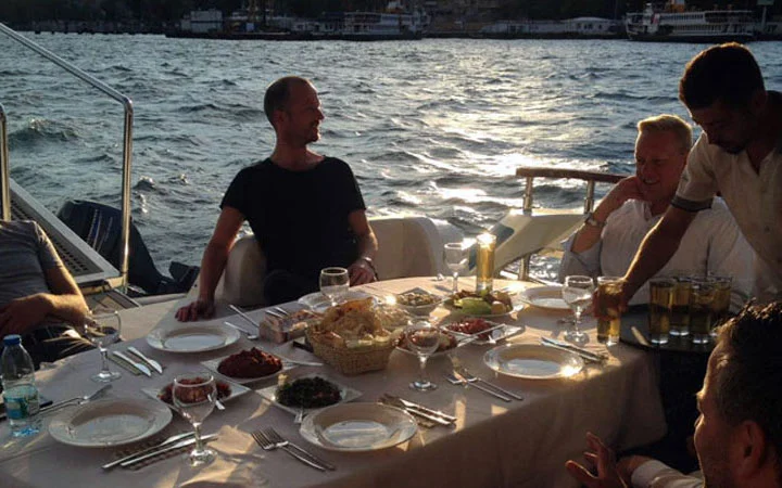Four people are sitting around a table set with dishes and drinks, dining outdoors on a private yacht with Iftar Cruise by the water in the evening sunlight. The mood is relaxed and cheerful, with delicious food and drinks on the table.