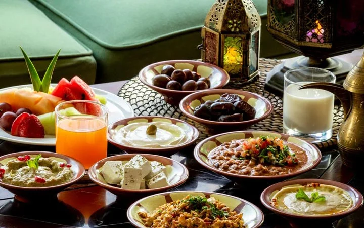 A table set with assorted Middle Eastern dishes, including hummus, labneh, fruit, dates, juice, and milk, surrounded by decorative lanterns—perfect for a Private Yacht with Iftar Cruise. The food is served in small bowls on a patterned mat.