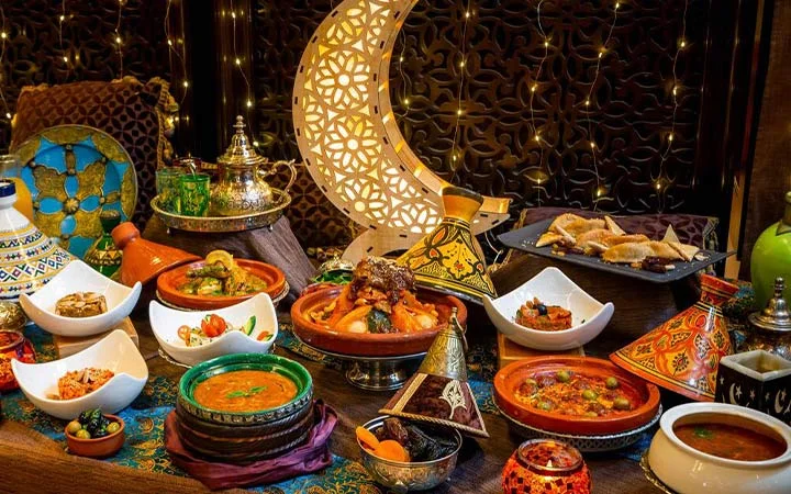 A colorful spread of Moroccan dishes, including tagines, soups, and bread, is arranged on a decorated table aboard a private yacht with Suhoor cruise, surrounded by ornate lanterns, pottery, and a golden crescent moon against twinkling lights.