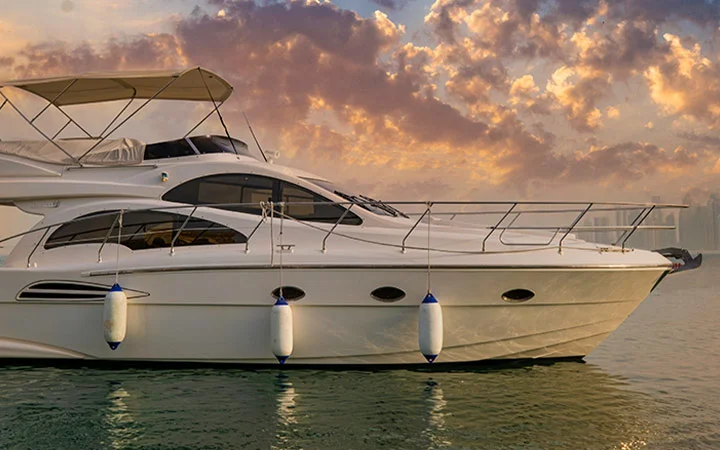 A white luxury private yacht with Suhoor cruise floats on calm water at sunset, beneath orange and purple clouds. The boat features blue and white fenders along its side and a shaded upper deck, perfect for a serene evening experience.