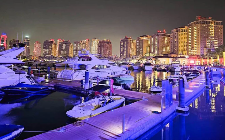 A marina at night with yachts and boats docked along illuminated piers, reflecting city lights from tall, modern buildings—a perfect setting for a private yacht with Suhoor cruise.