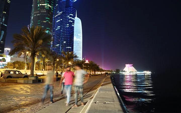 Nighttime cityscape along a waterfront promenade with modern skyscrapers, palm trees, and blurred figures enjoying a Ramadan 5K Walk and Karak With Guided Tour, colorful lights reflecting on the water beside parked cars.