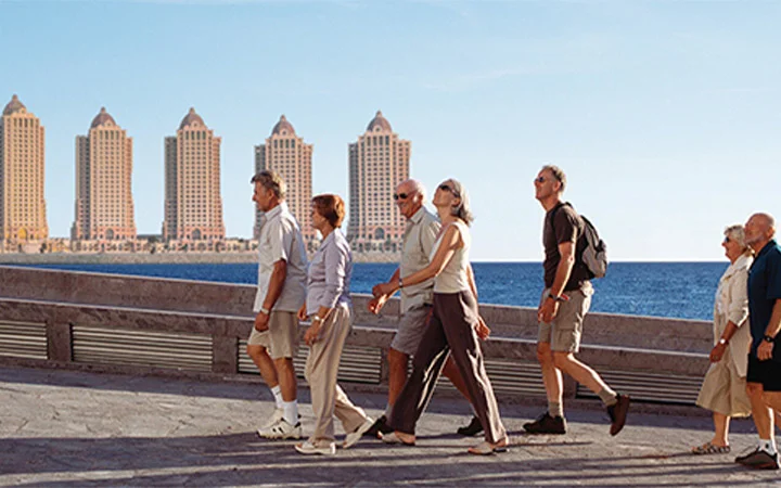 A group of people walk along a waterfront promenade under a clear sky, enjoying the Ramadan 5K Walk and Karak With Guided Tour. They appear relaxed and casually dressed, with tall buildings and the blue sea in the background.