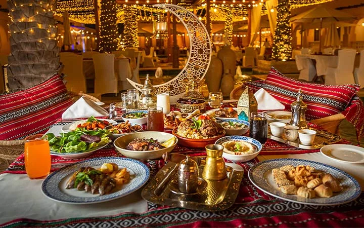 A festive outdoor dining table is set with various Middle Eastern dishes, drinks, and pastries for a Ramadan Souq Hunt & Suhoor. Decorative lights and a crescent moon centerpiece create a warm, celebratory atmosphere.