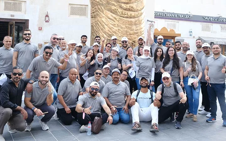 A large group in matching gray shirts pose and smile outdoors during the Ramadan Souq Hunt & Suhoor. Some give thumbs-up or peace signs, while a few hold bags; buildings and a golden sculpture are visible in the background.