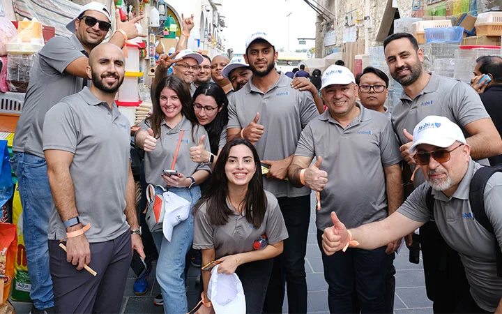 A group of people wearing matching gray polo shirts and white caps smile and give thumbs up while gathered closely together on a busy outdoor street during the Ramadan Souq Hunt. Some hold hats and phones, with colorful bins stacked nearby.