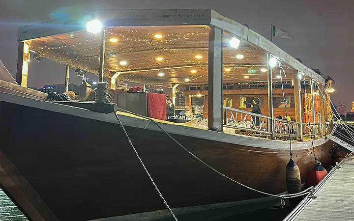 A wooden boat with string lights on its roof is docked at night for a Suhoor Cruise. The warmly lit interior reveals tables and chairs, creating a cozy atmosphere as the boat rests tied to the pier with ropes.