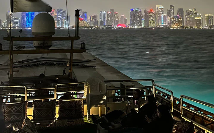 A nighttime view from a Suhoor Cruise boat deck with people sitting on patterned cushions, gazing at a brightly lit city skyline across the water. The illuminated buildings shimmer in various colors, creating a magical atmosphere.