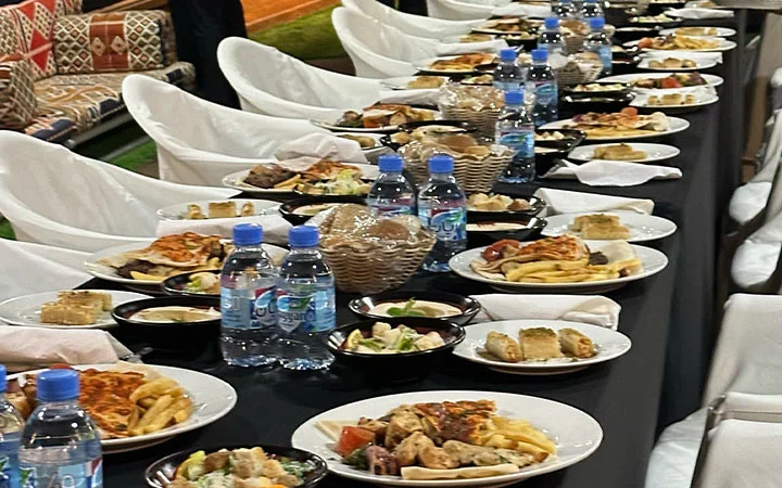 A long table is set with plates of assorted food, bottled water, salads, bread baskets, and napkins. Surrounded by white chairs, it suggests a lively Suhoor Cruise banquet or large gathering.