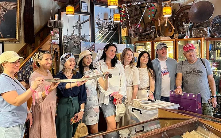 A group of nine people poses inside an antique or curiosity shop filled with vintage items, animal mounts, and framed photos. Smiling, they gather around a woman in the center holding a long, curved sword—ready for an Eid Treasure Hunt adventure.