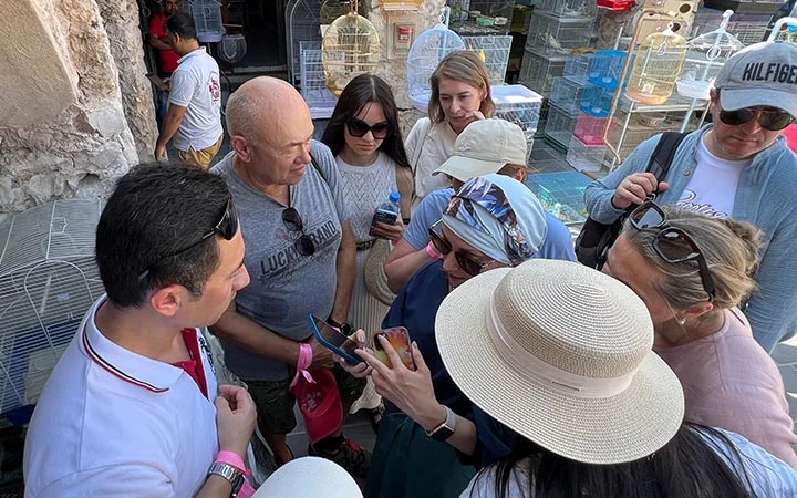 A group of people stand closely together outdoors, looking at a phone held by a woman in a sunhat. Birdcages in the background suggest a market setting, where the group appears engaged—possibly taking part in an Eid Treasure Hunt.