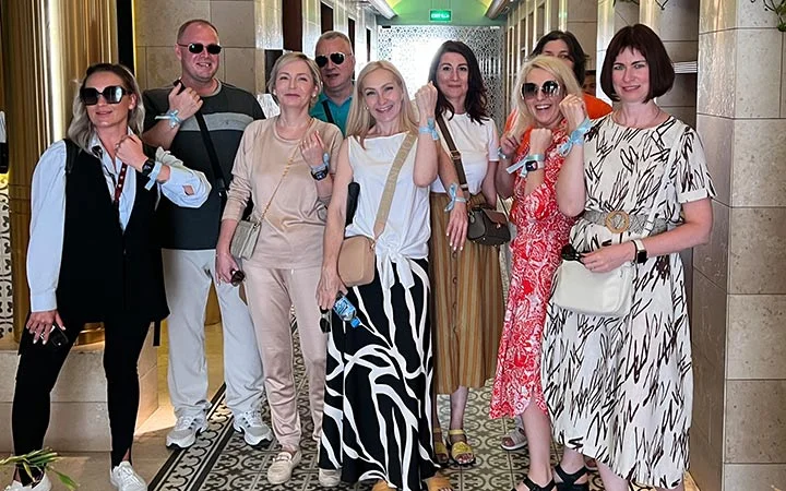 A group of eight adults, five women and three men, stand indoors smiling at the camera. Casually dressed with sunglasses and wristbands, they appear to be enjoying an Eid Treasure Hunt social gathering.