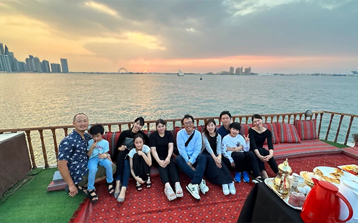 A group of ten people sits together on a boat deck at sunset, celebrating Eid-Swim and Jump. With smiles and relaxed vibes, they're surrounded by red cushions and a table set with food and a teapot, city skyline and calm water in the background.