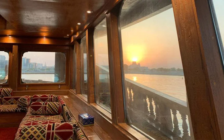 Interior of a wooden boat with patterned seating, large windows showing a sunset over a city skyline and river, creating a warm and cozy atmosphere—perfect for an Eid-Swim celebration or spontaneous jump into adventure.