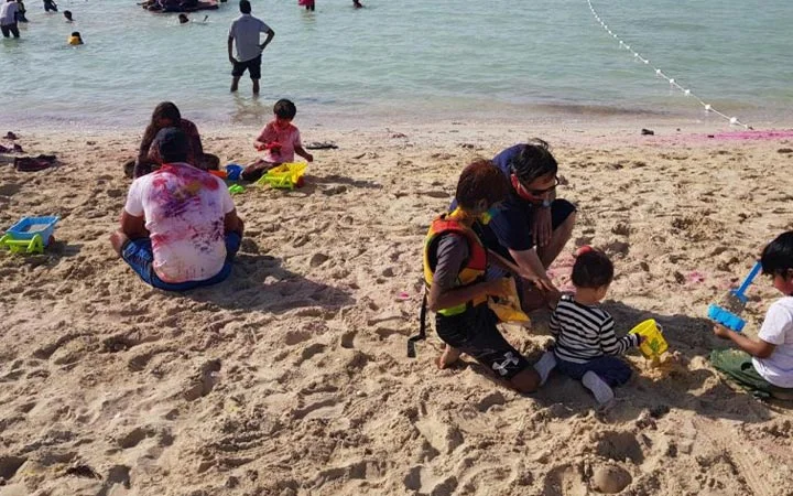 Children and adults play in the sand and swim at a beach, enjoying an Eid-Swim under sunny skies. Some build sandcastles with buckets and toys, while others jump into the water on this warm day.